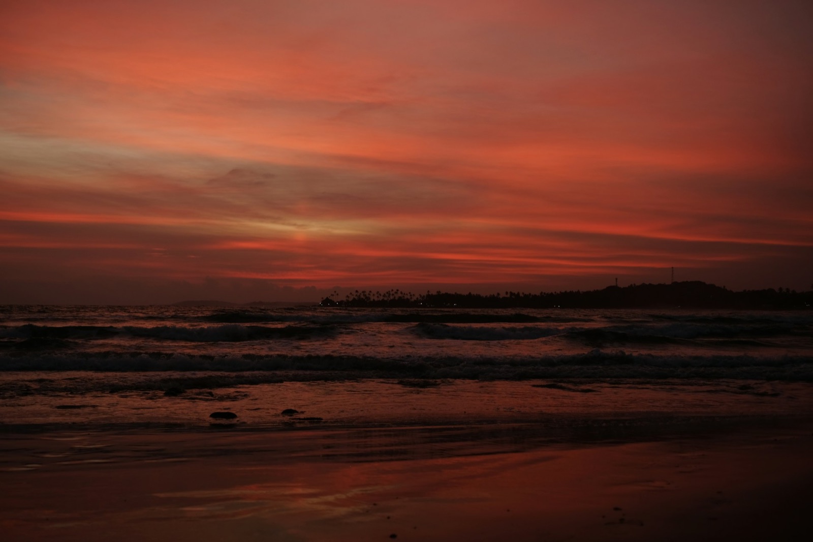 A red sunset over waves and a distant palm-lined coast at Dickwella Beach, Sri Lanka.