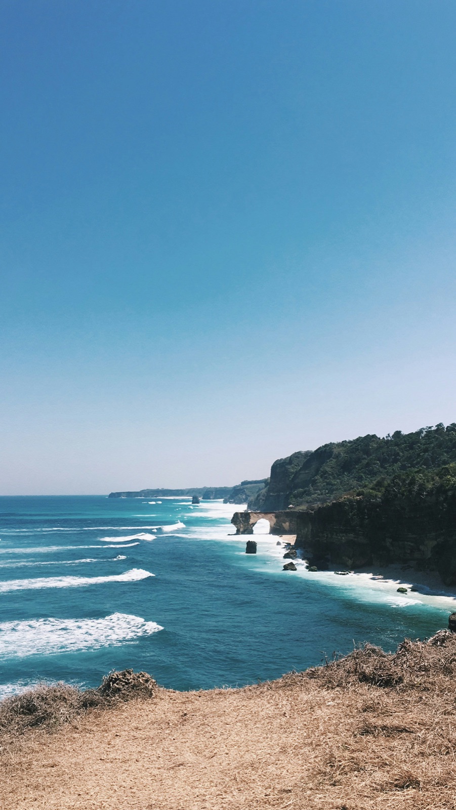 Turquoise water and white surf along a Sumba coast, with green hills in the foreground.